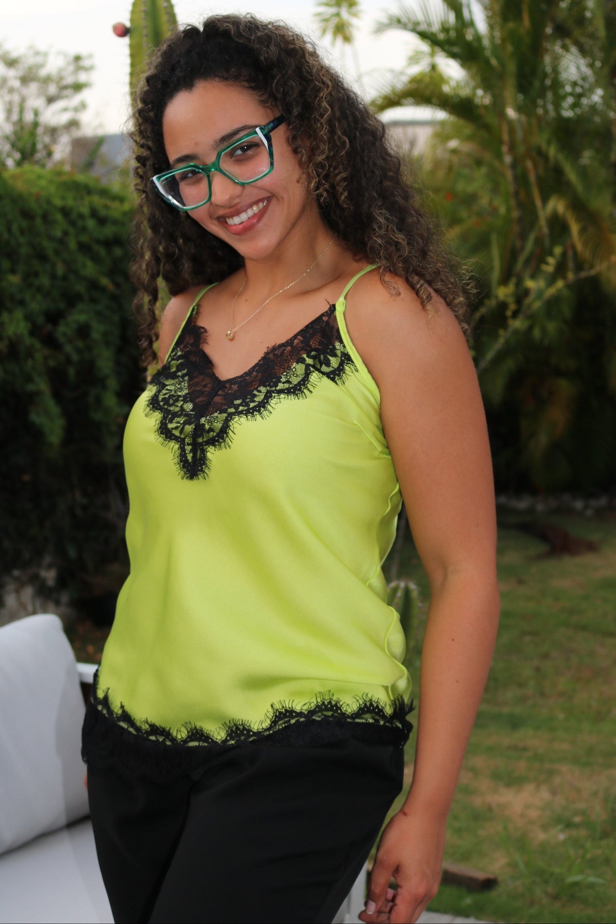 A woman wearing a bright lime green sleeveless top with black lace trim and black pants, photographed outdoors on a patio with palm trees and grass in the background.
