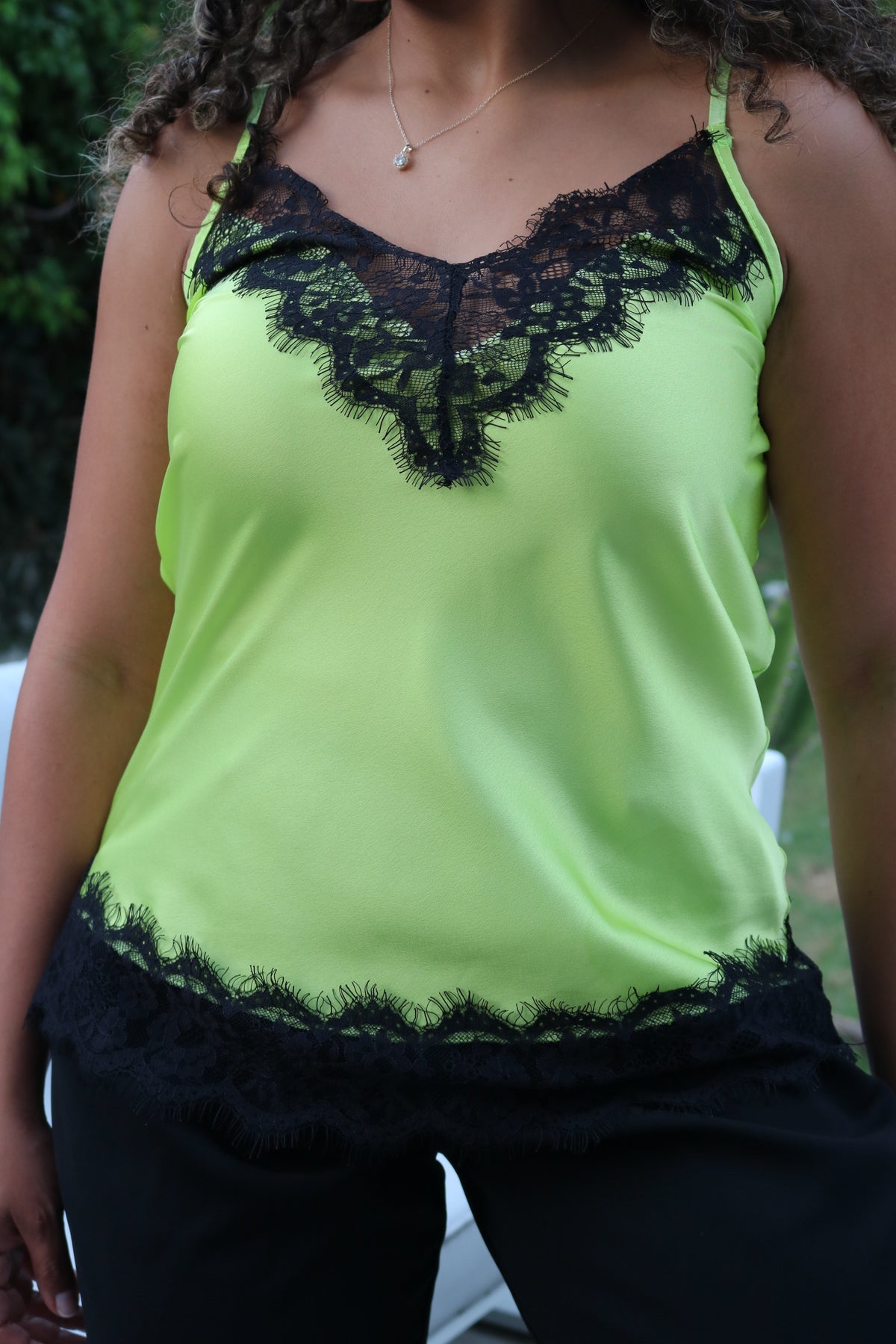 A close-up of a person wearing a neon yellow-green camisole top with black lace trim along the neckline and hem, paired with black bottoms.